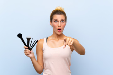 Young blonde woman holding lots of makeup brush surprised and pointing front