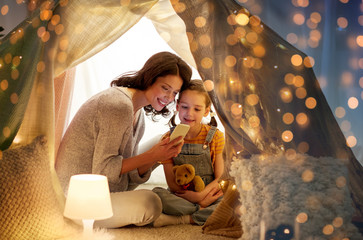 Sticker - family, hygge and technology concept - happy mother and little daughter with smartphone in kids tent at night at home