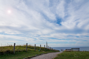 Landscape with road with two people in the background and two benches next to the coast of Comillas in Cantabria, Spain