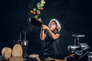 Wall Mural - Funny little girl is tossing vegetables on the pan at dark photo studio.