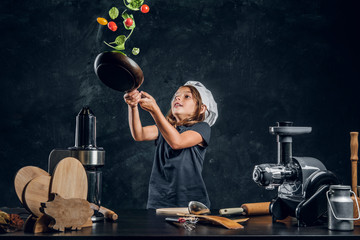 Wall Mural - Cheerful girl is tossing vegetables on the pan at dark photo studio.