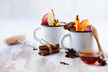 Two white metal mugs with ingredients for hot mulled wine - traditional christmas beverage. Ripe apple and orange, spices and honey. Corkscrew to open wine bottle. Holiday festive mood, rustic style