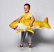 © FAB.1 - little boy, dressed in festive clothes, wears a balloon in the shape of a yellow shark fish, celebrates a holiday, has a wide smile, stands on a light background, being in a good mood.