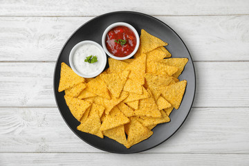 Black plate with tasty Mexican nachos chips and sauces on white wooden table, top view