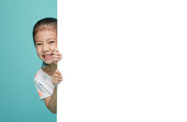 Smiling happy cute child hiding behind a blank white board, empty space in studio shot isolated on colorful blue background