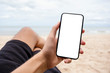 © Farknot Architect - Mockup image of a hand holding and showing black mobile phone with blank desktop screen while sitting on the beach chair