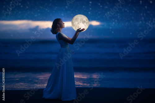 Photographie A young girl on a night beach holds the moon, with a starry sky