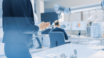 Wall Mural - Stylish Female Industrial Robotics Engineer Uses Tablet Computer with CAD Software and to Manipulate and Program Robot Arm for Lifting, Moving Objects. Technology Research Facility for Cloud Computing