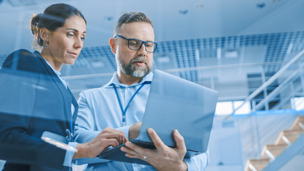 Poster - Stylish Confident Businesswoman and Businessman Working on a Project, Man Holds Laptop Computer, They Reference it. In the Background Modern and Bright Facility. Low Angle Shot