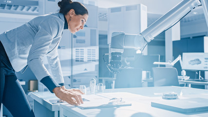Leinwandbilder - Stylish Female Robotics Engineer Leans on the Table Works with Blueprints, Documents and Tablet Computer, She's Programming Robot Arm Movements