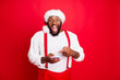 Portrait of impressed black santa claus in xmas hat pointing at his cell phone device screaming wow omg got like wearing white pullover isolated over red background