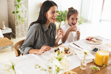 Wall Mural - image of smiling family mother and little daughter eating together while having breakfast at home in