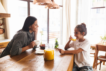 Wall Mural - image of positive family mother and little daughter smiling and having breakfast at home in morning