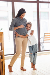 Wall Mural - full length image of joyful family little girl smiling and hugging her pregnant mother at cozy apart