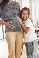 Wall Mural - image of joyful excited little girl smiling and hugging her pregnant mother's belly at cozy apartmen