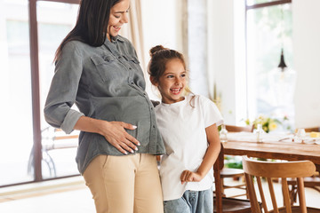 Wall Mural - image of adorable family little girl smiling and hugging her pregnant mother иat cozy apartment