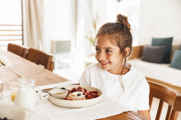 Wall Mural - Portrait of happy little girl sitting at table in bright kitchen while having breakfast