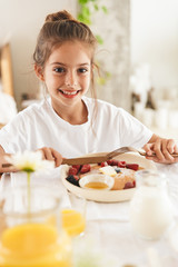 Wall Mural - portrait of joyful little girl sitting at table in bright kitchen while eating pancakes with raspber