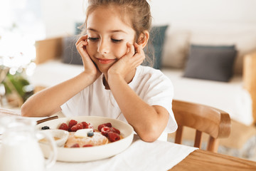 Wall Mural - portrait of cute little girl sitting at table in bright kitchen at home while having breakfast