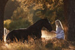 portrait of beautiful young woman with blond hair sitting in front of lying horse and feed it from hands in sunset sunlight in autumn