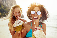 Two Laughing Friends Drinking From Coconuts On A Sandy Beach