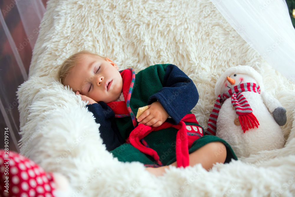 Baby boy, cute child, wearing santa claus robe sitting in rocking chair with Christmas tree and lights on