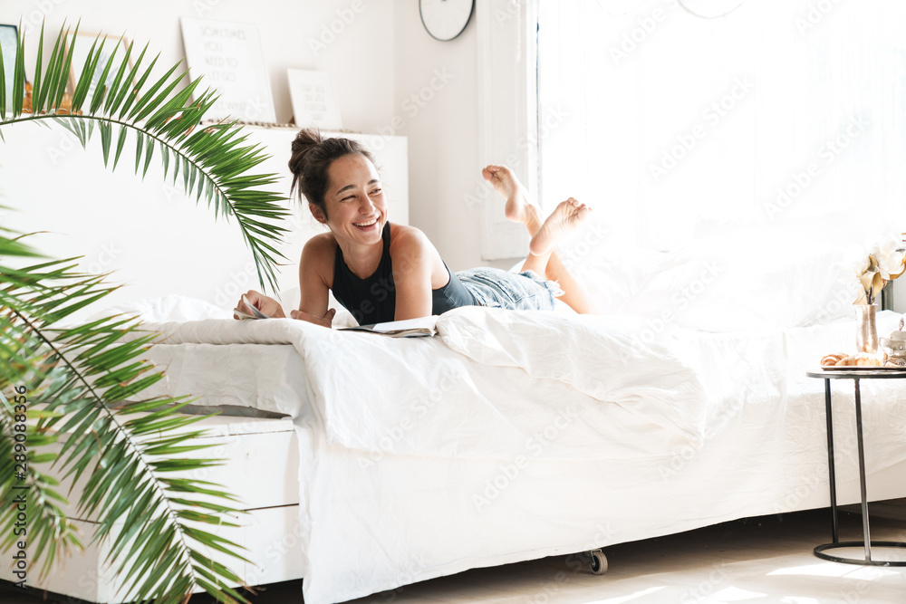 Portrait of pretty woman smiling while lying and reading magazine on bed in bright room with green plant - obrazy, fototapety, plakaty 