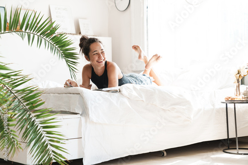 Cuadros en Lienzo Portrait of pretty woman smiling while lying and reading magazine on bed in brig