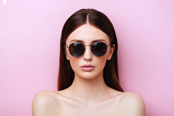 Wall Mural - Portrait of an attractive young girl in round glasses on a pink background in studio