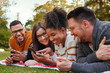 © StratfordProductions - Smiling mixed race group of friends lying together on green grass using mobile phone in the park - friends having a picnic
