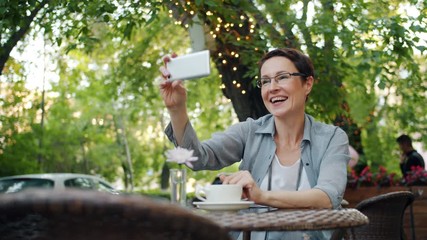 Wall Mural - Mature lady in glasses is making online video call relaxing in street cafe in city sitting at table alone talking holding smartphone. Conversation and internet concept.