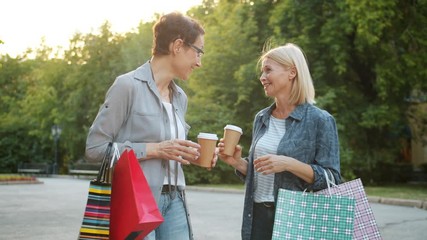 Wall Mural - Slow motion of beautiful mature women chatting holding shopping bags and coffee walking outside in park. People, lifestyle and consumerism concept.