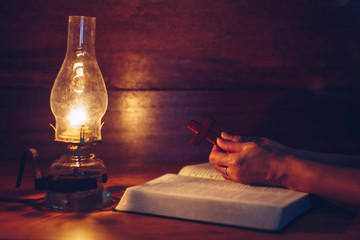 Wall Mural - Close up of woman hands hold wooden cross over open bible with oil lamp on wood table while reading bible in the dark room, Christian background devotion or bible study concept
