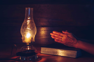 Wall Mural - Close up of woman hands praying on bible with oil lamp on wooden table in dark room, Christian faith concept background