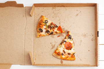 Closeup open box with two slices of pizza with pepperoni, minced meat, mushrooms, sweet pepper and olives on a white wooden table. Top view.