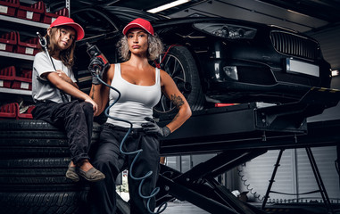 Experienced serious woman and her little helper are posing for photographer at dark auto service as great team.