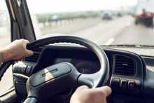 Male Driver Hands Are Holding Steering Wheel Of Truck During The Movement In The Road. Image With Selective Focus On The Wheel, Dashboard And Blurred Windshield