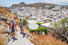 A Man And Two Women Stand On A Path In The Background Of The City Of Lindos .Horizontally.