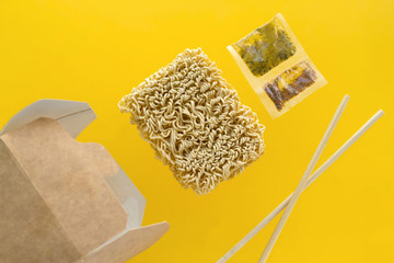 flying on a yellow background cardboard box, wooden sticks, dry instant noodles and a packet of spices, Asian fast food levitating on a yellow background