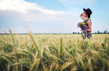 Wall Mural - Cute attractive girl with a bouquet of colorful flowers in her hands. Young woman breathes in the scent of plants on wheat field during sunset. Pensive look. Romantic atmosphere.