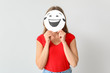 © Pixel-Shot - Young woman hiding face behind emoticon against light background