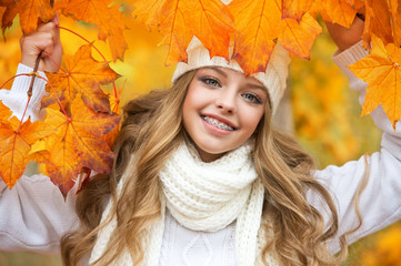 Portrait of beautiful young woman walking outdoors in autumn. Girl with autumn yellow leaf in knitted hat and sweater.  Portrait of a girl with orthodontic appliance.