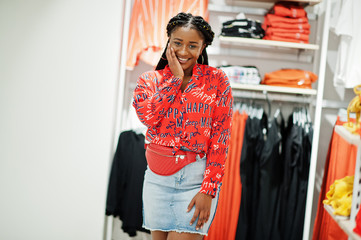 Wall Mural - Modish african american woman in red shirt, jeans skirt with crossbody bag posed at clothes store. It's time for shopping.