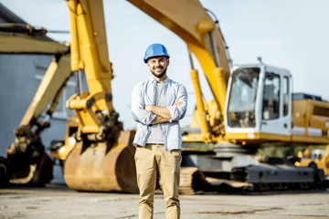 Portrait of a handsome builder standing on the open ground of the shop with heavy machinery for construction