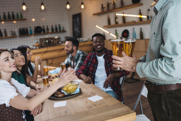 Wall Mural - cropped view of man holding glasses of light beer near multicultural friends in pub