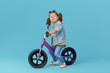 © oes - Little girl learns to keep balance while riding a bicycle. Stylish child in sunglasses  isolated on blue background