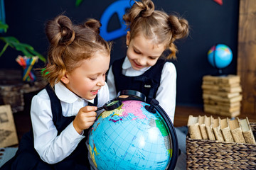 little twin girls in school uniform using magnifying glass to examine globe while sitting on floor d