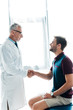 © LIGHTFIELD STUDIOS - happy doctor in glasses shaking hands with cheerful young man in clinic