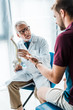 © LIGHTFIELD STUDIOS - selective focus of bearded doctor in glasses holding spine model near patient gesturing in clinic
