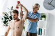 chiropractor in glasses standing near man exercising with dumbbell in clinic
